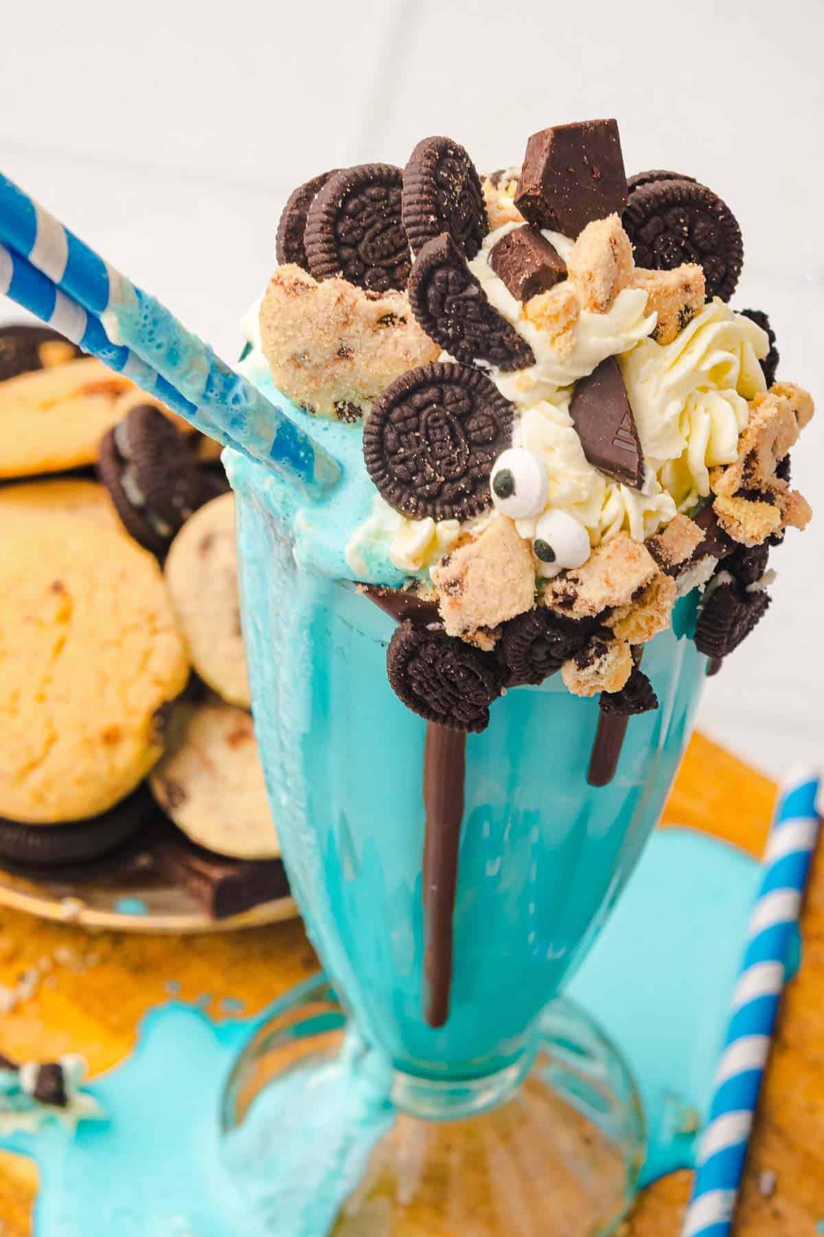 A Cookie Monster Milkshake topped with whipped cream, mini cookies, chocolate pieces, and candy eyes, served with two straws&mdash;a fun and delicious Milkshake Recipe!.