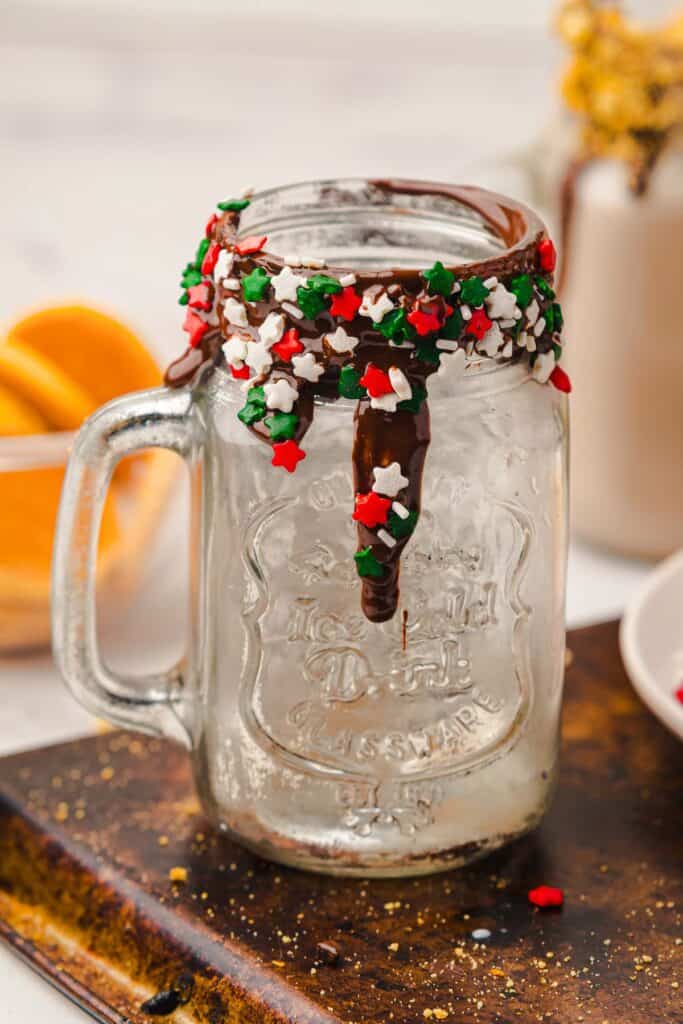 A mason jar, perfect for a Gingerbread Freakshake, is rimmed with chocolate and festive red, green, and white star sprinkles, sitting on a wooden board.