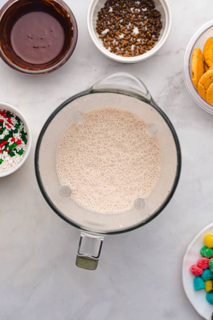 A blender with a frothy liquid sits on a white surface, surrounded by bowls of sprinkles, cookies, and chocolate&mdash;perfect inspiration for crafting a festive Christmas recipe or a decadent Gingerbread freakshake.