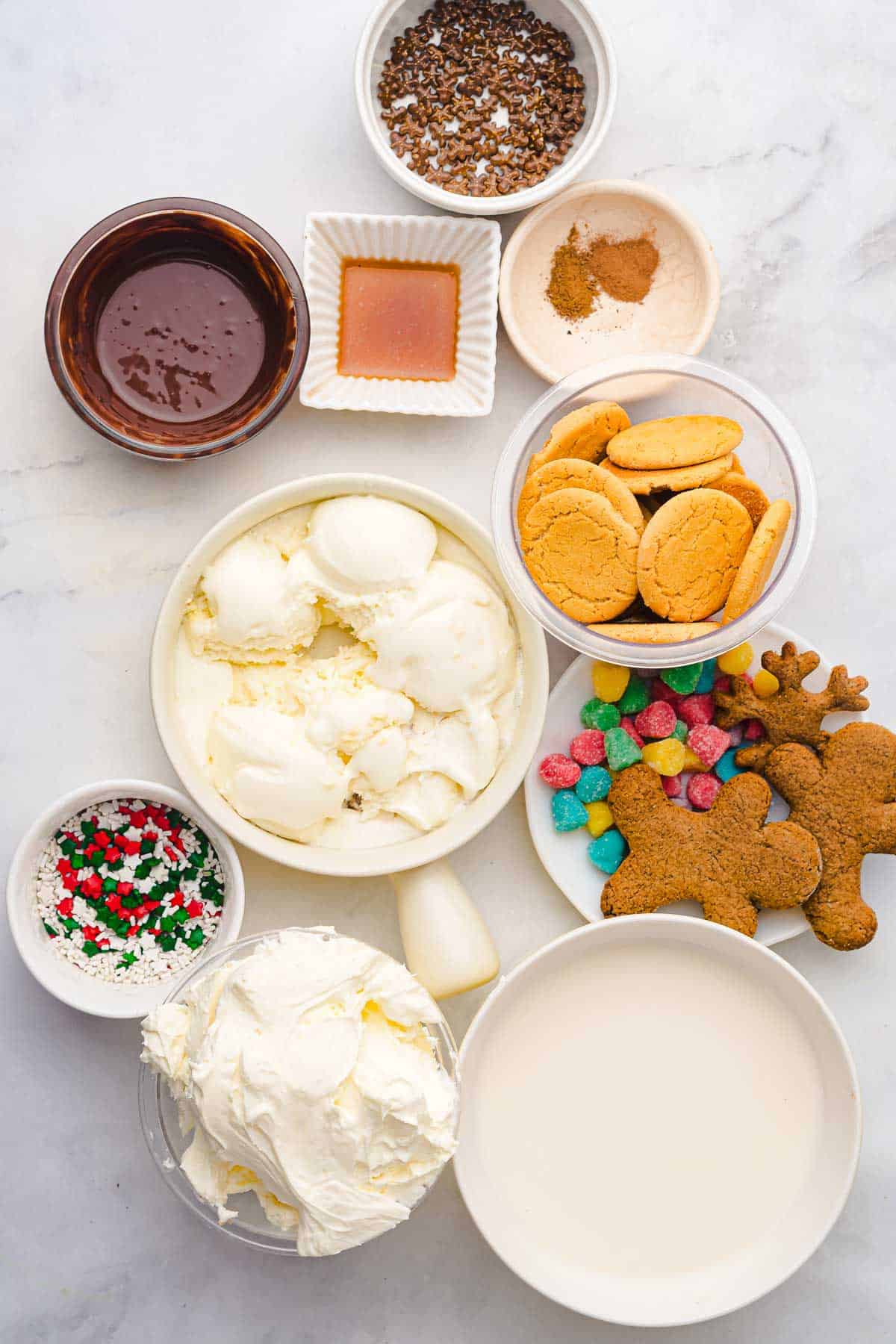 Various bowls with ice cream, cookies, candies, sprinkles, chocolate sauce, and whipped cream on a white surface&mdash;perfect ingredients for building your own gingerbread freakshake masterpiece.