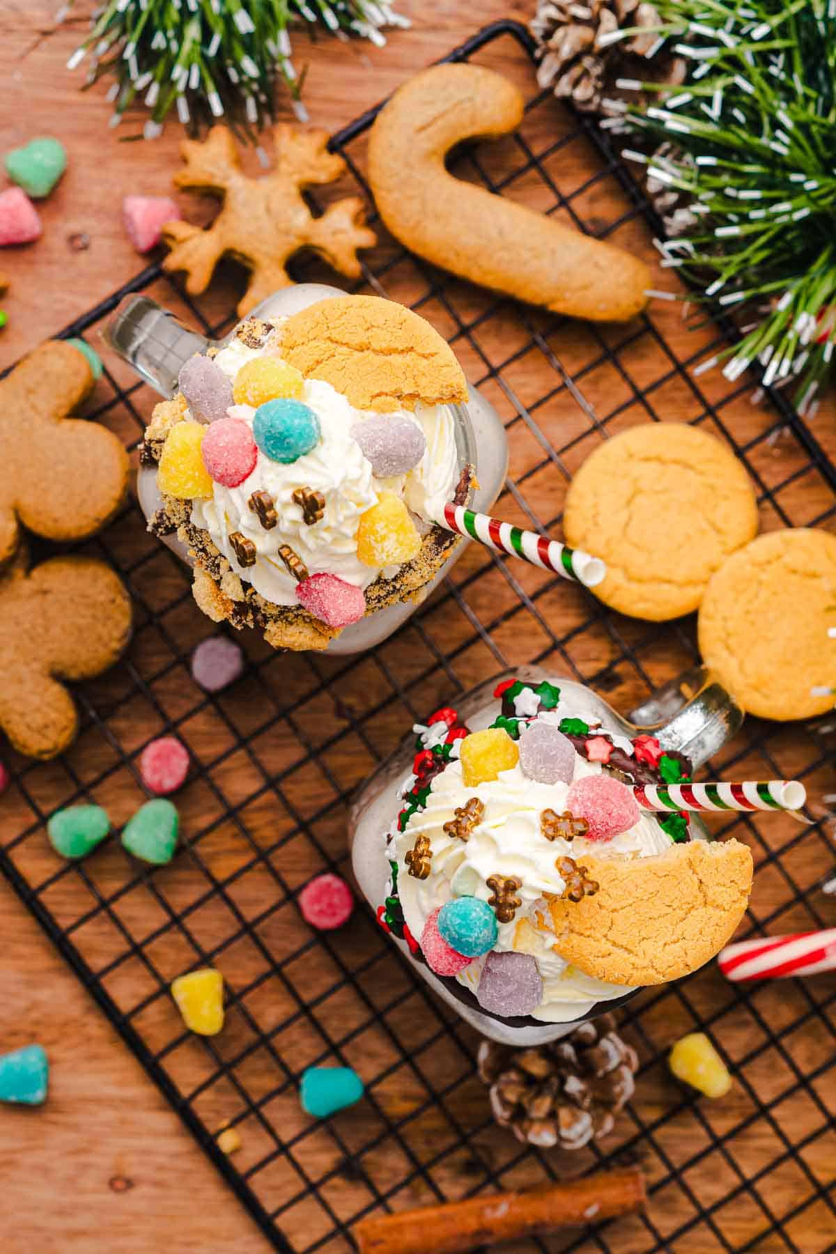 Two festive mugs of holiday milkshakes, inspired by a Gingerbread Freakshake, are topped with whipped cream, cookies, sprinkles, and gumdrops on a wire rack. Perfect for trying out your next Christmas Recipe!.
