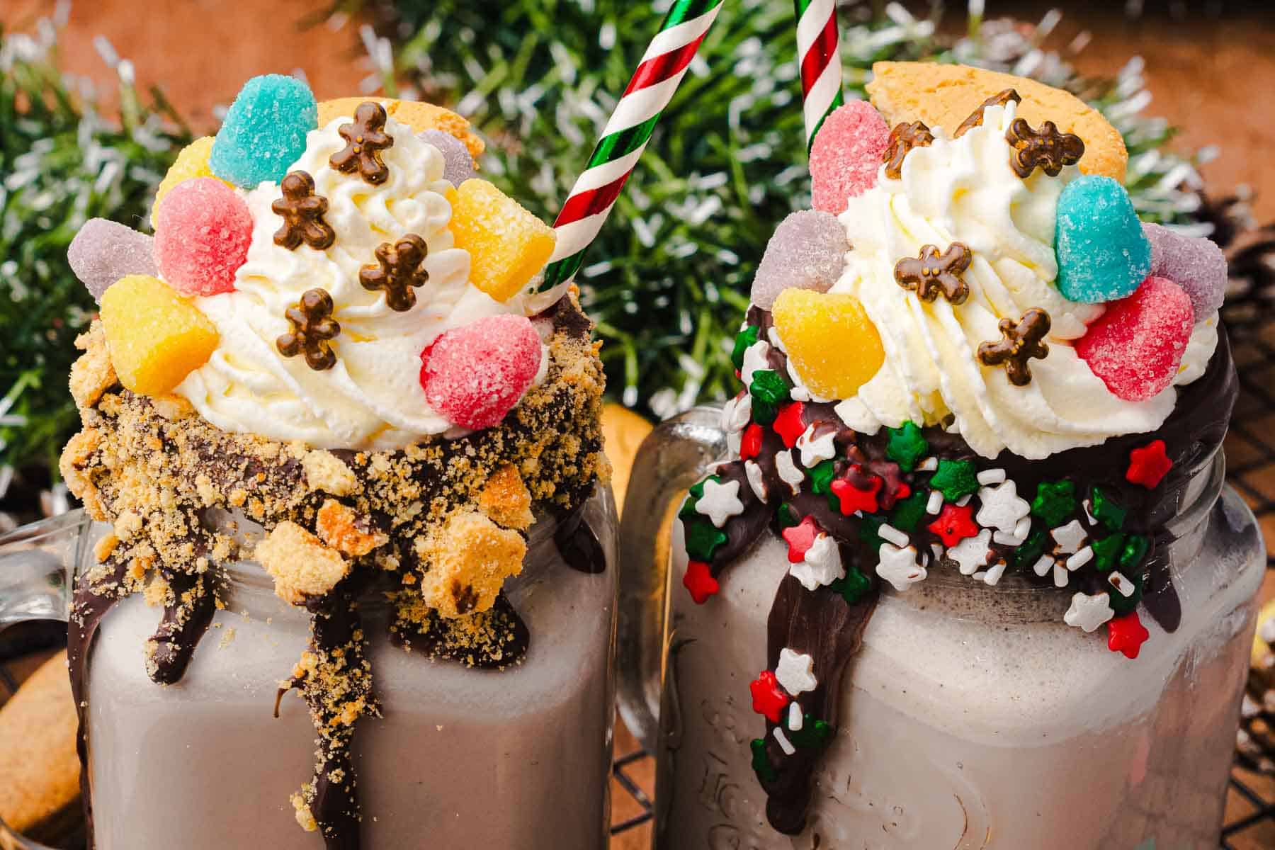 Two festive Gingerbread Freakshakes in mason jars, topped with whipped cream, candies, sprinkles, and striped straws&mdash;perfect for a Christmas treat.