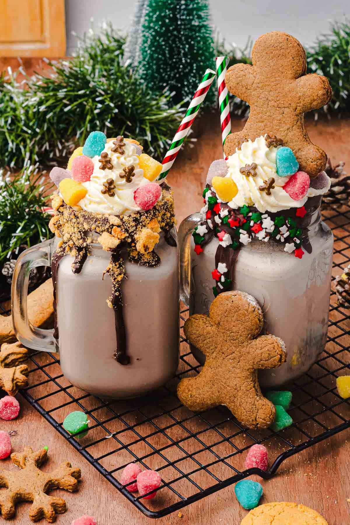Two festive mugs of hot chocolate topped with whipped cream, candies, and gingerbread cookies on a cooling rack&mdash;perfect for a cozy Christmas Recipe or your next Gingerbread Freakshake craving.