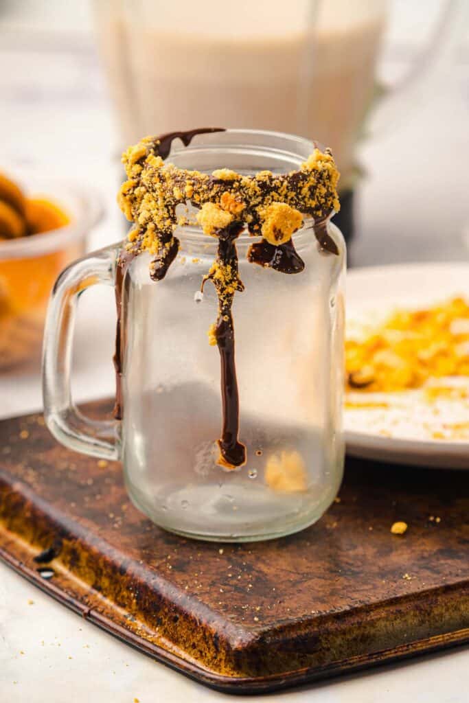 A mason jar mug with chocolate and crumbs on the rim holds a delightful Gingerbread Freakshake, sitting on a tray with plates and snacks nearby.
