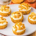 Six mini gingerbread cheesecakes with cream cheese frosting and crumbled cookie pieces on top are arranged on a white plate. A whisk, a wooden spoon, a gingersnap cookie, and a glass of milk are in the background.