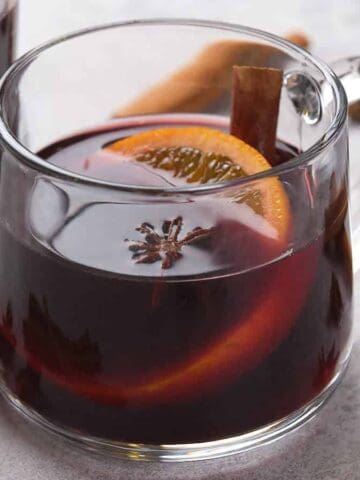 Two clear glass mugs of mulled wine with orange slices, star anise, and cinnamon sticks.