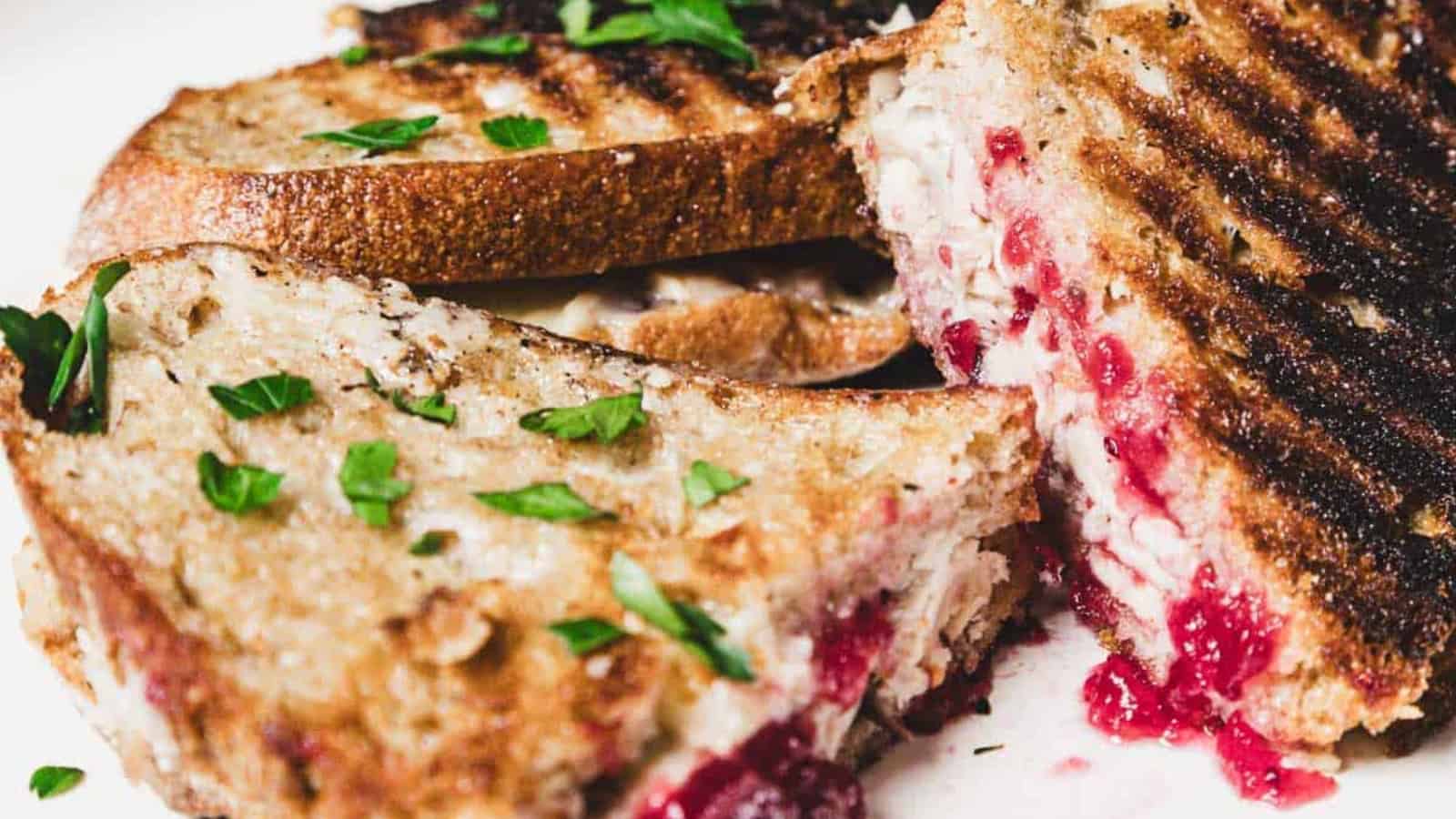 Grilled turkey and cranberry sandwiches with fresh herbs sprinkled on top, sliced and served on a white plate.