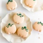 Five pumpkin-shaped macarons with green icing stems on white plates, viewed from above.