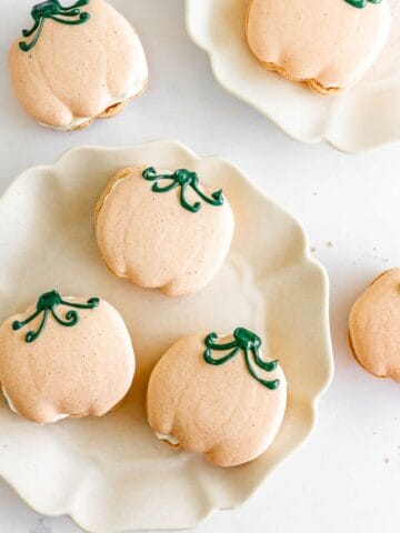 Five pumpkin-shaped macarons with green icing stems on white plates, viewed from above.