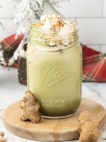 Iced Matcha Latte with whipped cream in a mason jar, topped with cinnamon and served alongside delicious gingerbread cookies.