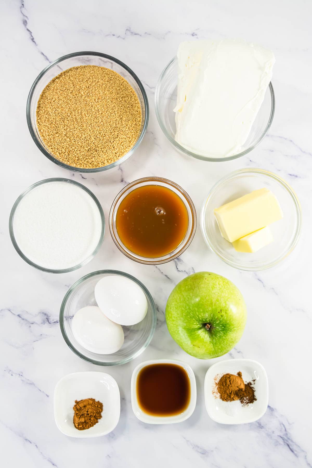 Bowl of graham crumbs, cream cheese, sugar, caramel, butter, eggs, green apple, spices, and vanilla on marble.