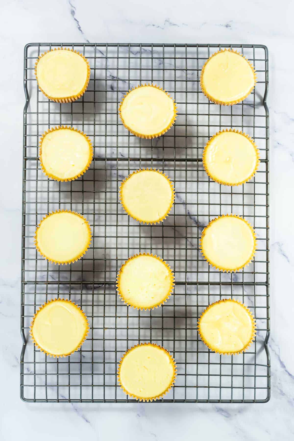 Twelve mini cheesecakes in yellow wrappers cooling on a wire rack over a marble surface.