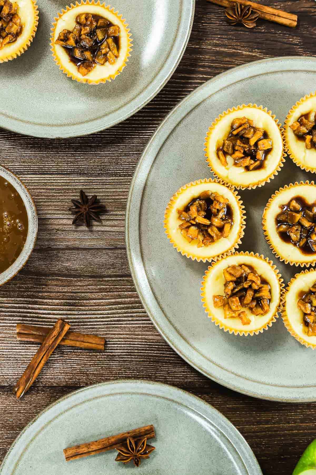 Mini cheesecakes topped with diced spiced apples on grey plates, with cinnamon sticks and star anise nearby.