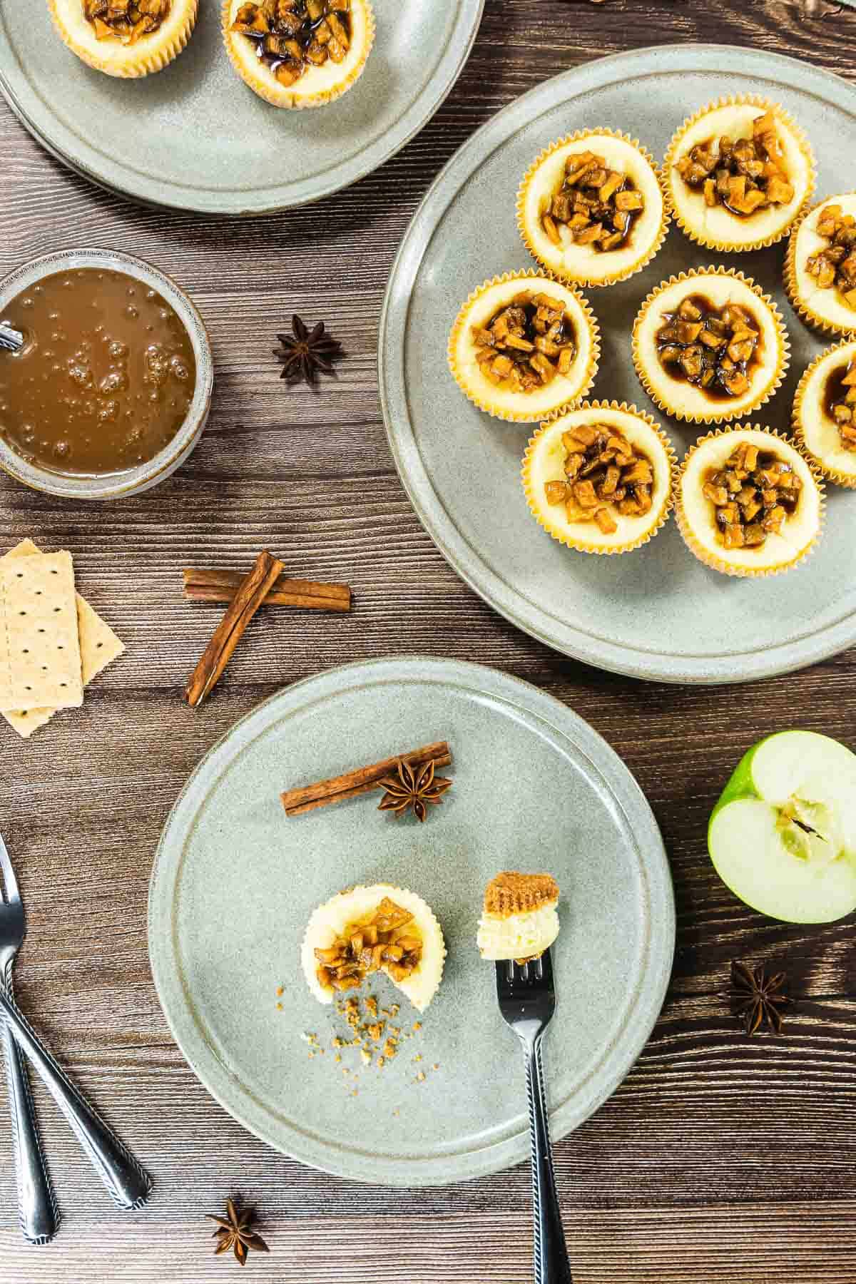 Mini apple pies on gray plates with spices, graham crackers, and a sliced green apple on a wooden table.