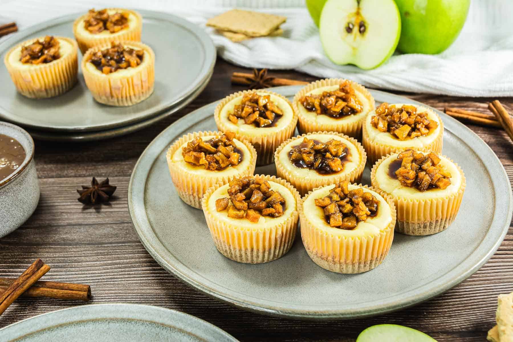 Mini cheesecakes topped with caramelized apple pieces on a plate, with green apples and cinnamon sticks nearby.