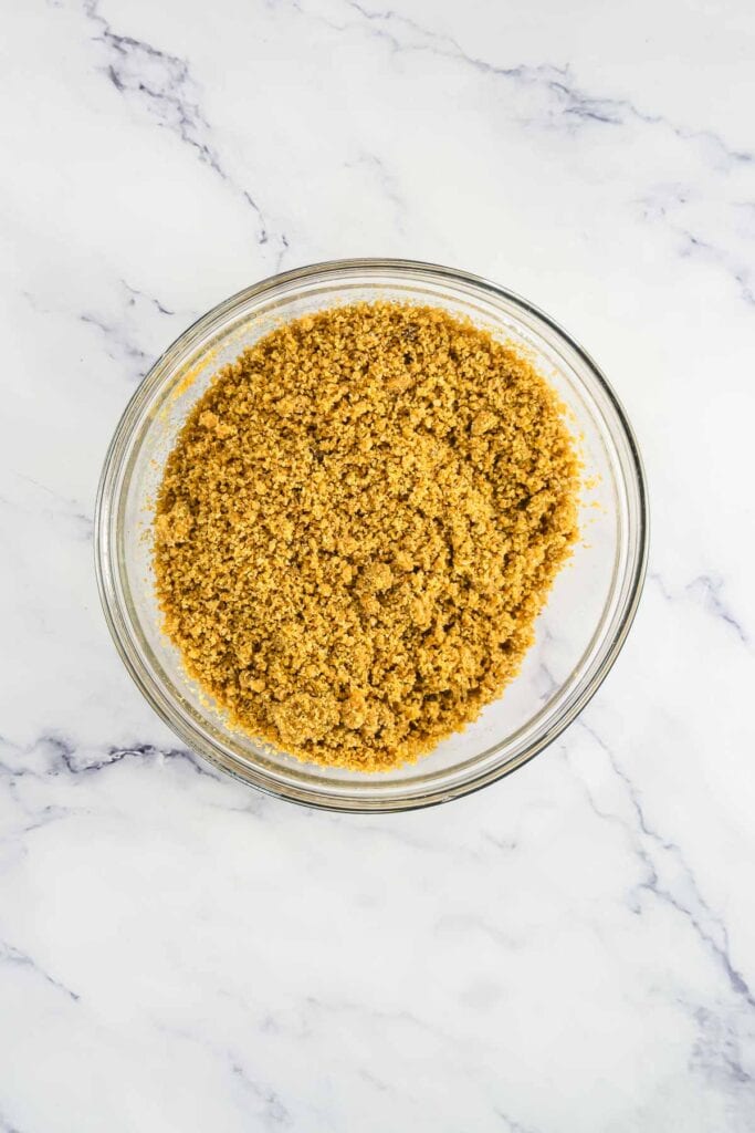 Glass bowl filled with crumbly graham cracker crust mixture on a white marble surface.