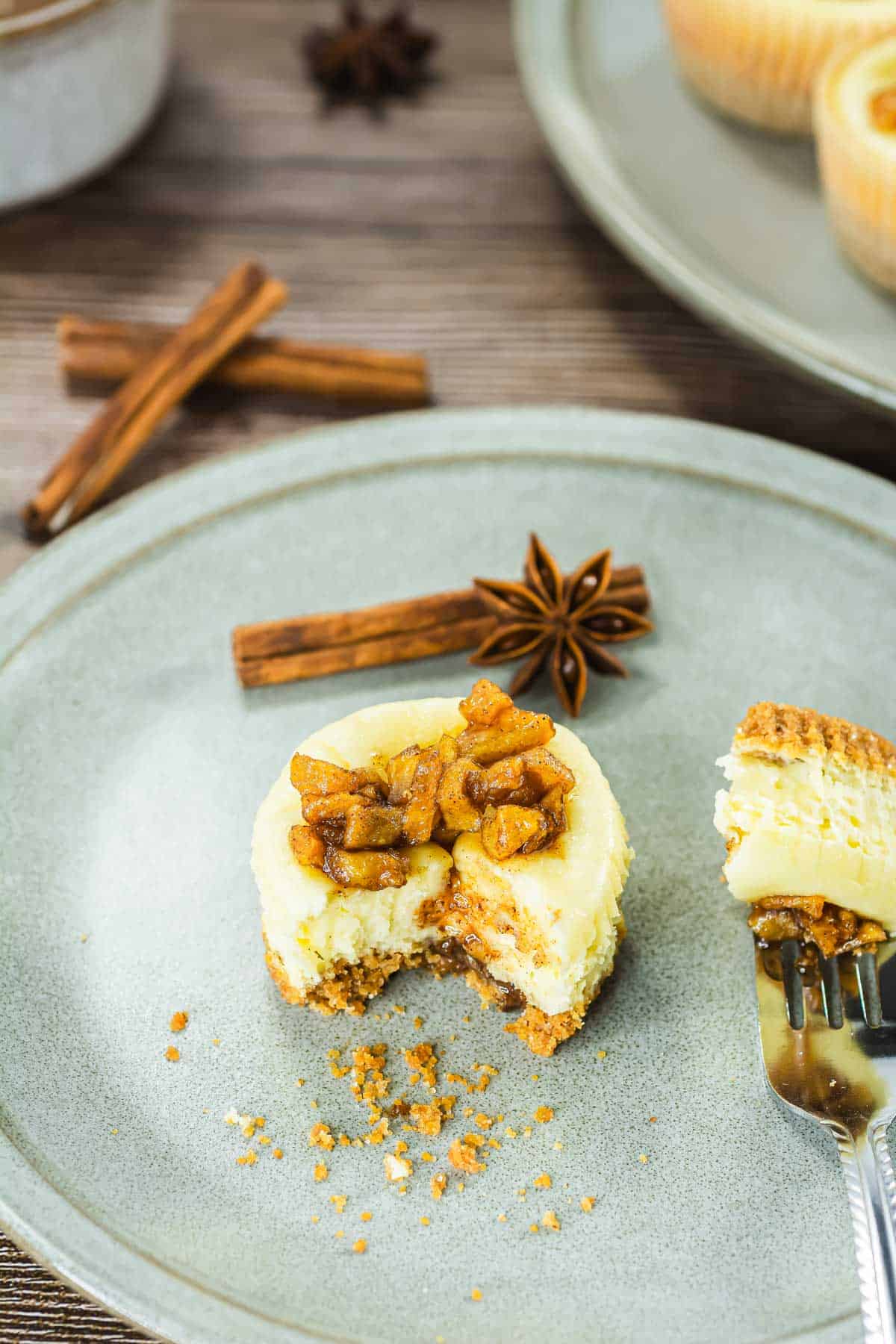 Mini cheesecake topped with spiced apple pieces on a gray plate, with cinnamon sticks and star anise nearby.