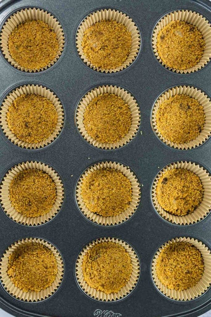 A muffin tin filled with paper liners, each holding a pressed crumb crust mixture.