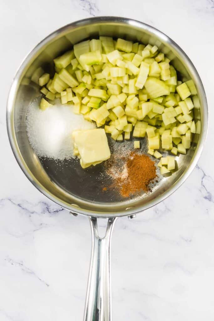 Diced apples, sugar, butter, and cinnamon in a saucepan on a marble countertop.