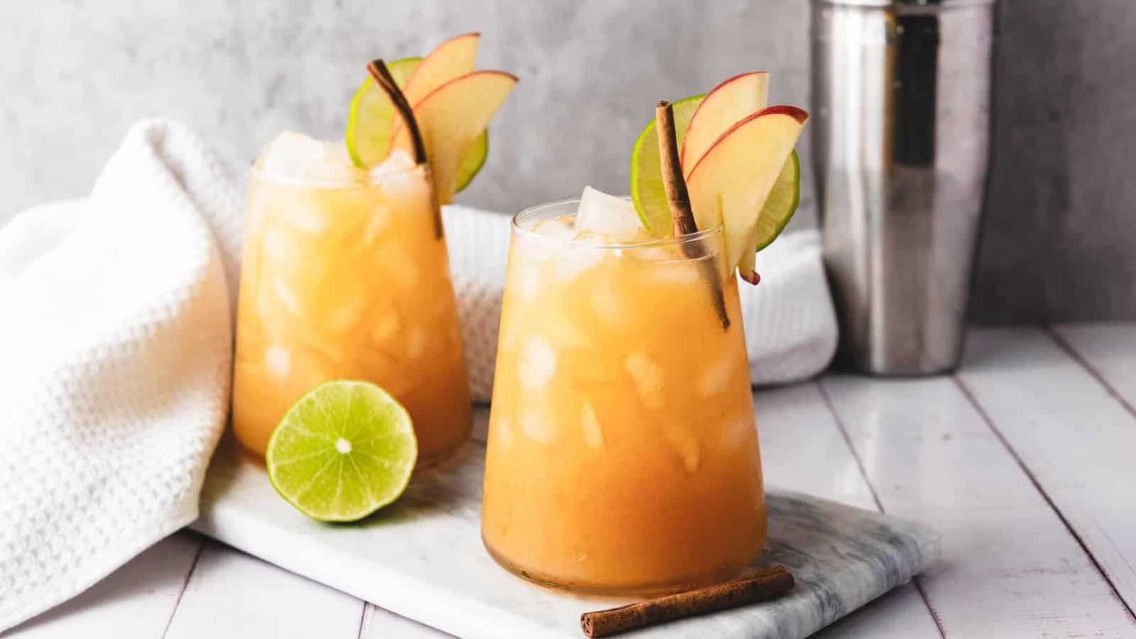 Iced apple cider drink garnished with apple slices, a lime wheel, and a cinnamon stick on a marble tray.
