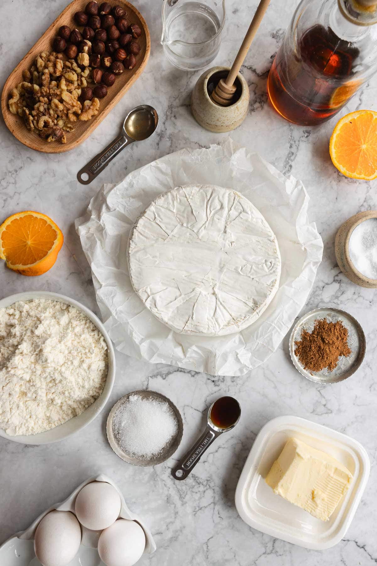 Round brie cheese on parchment surrounded by baking ingredients, nuts, orange halves, eggs, butter, and Puff Pastry on marble&mdash;perfect for crafting a festive Cranberry Brie Pastry.