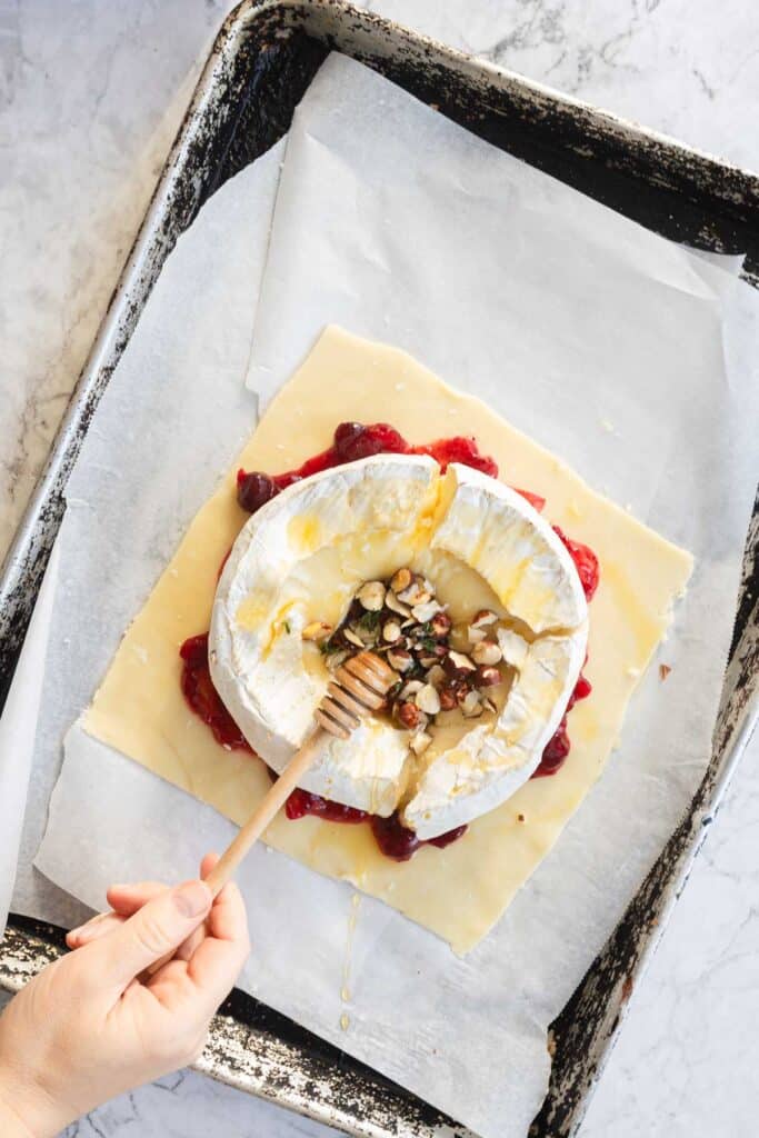 Hand drizzling honey over a wheel of Cranberry Brie on pastry with nuts and fruit, all arranged on a parchment-lined baking sheet&mdash;perfect for making Cranberry Brie Puff Pastry.