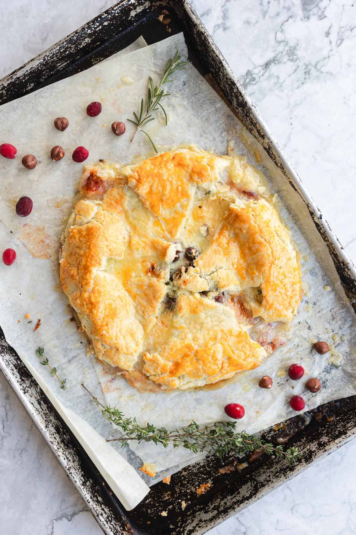 A round Cranberry Brie Puff Pastry on parchment paper, garnished with cranberries and herbs, sits on a rustic baking tray.