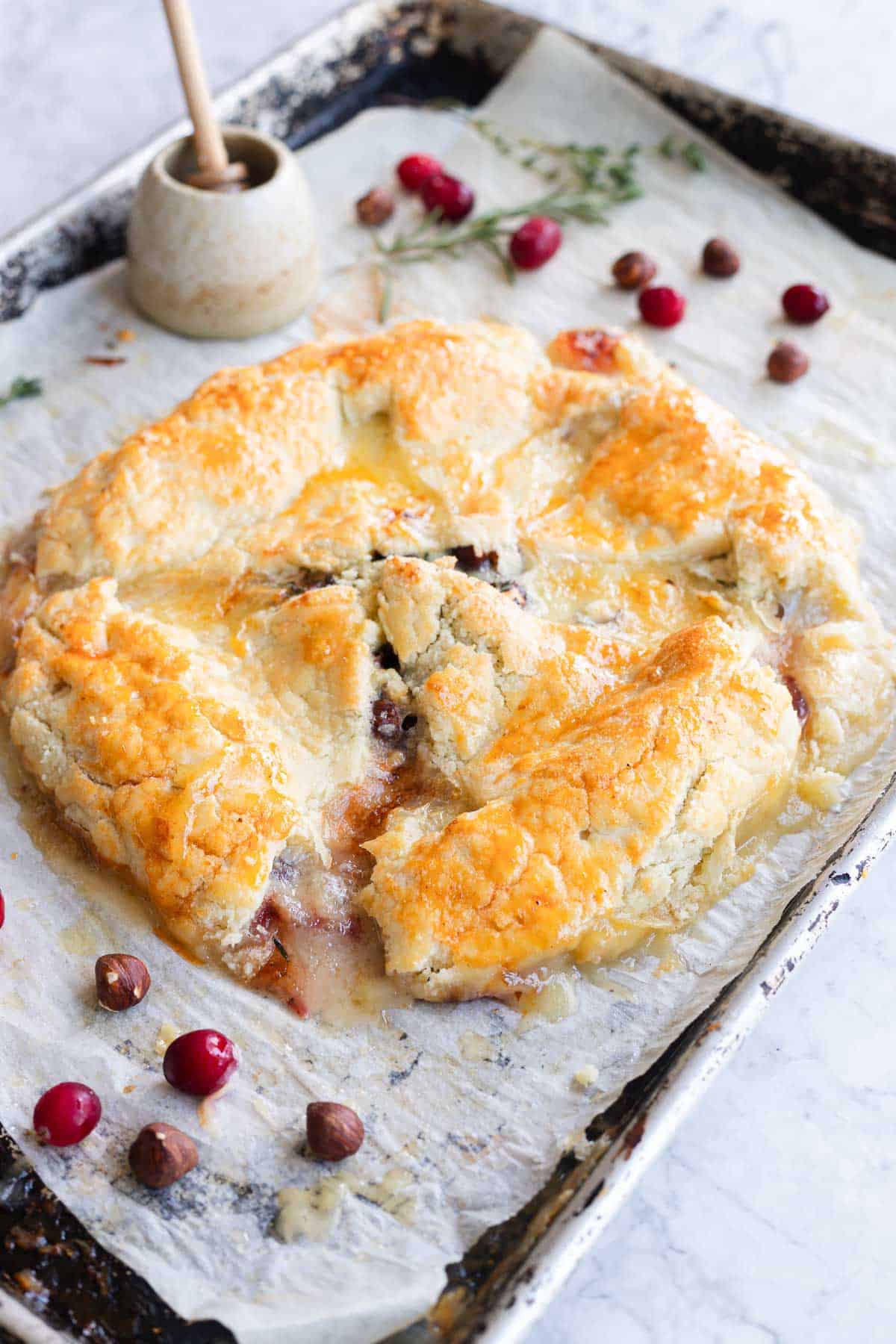 Golden-brown Cranberry Brie Puff Pastry with visible filling rests on parchment, surrounded by cranberries and hazelnuts.