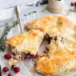 Golden cranberry galette with a slice being served, surrounded by cranberries and herbs on parchment paper—a rustic twist on a classic Cranberry Brie appetizer.