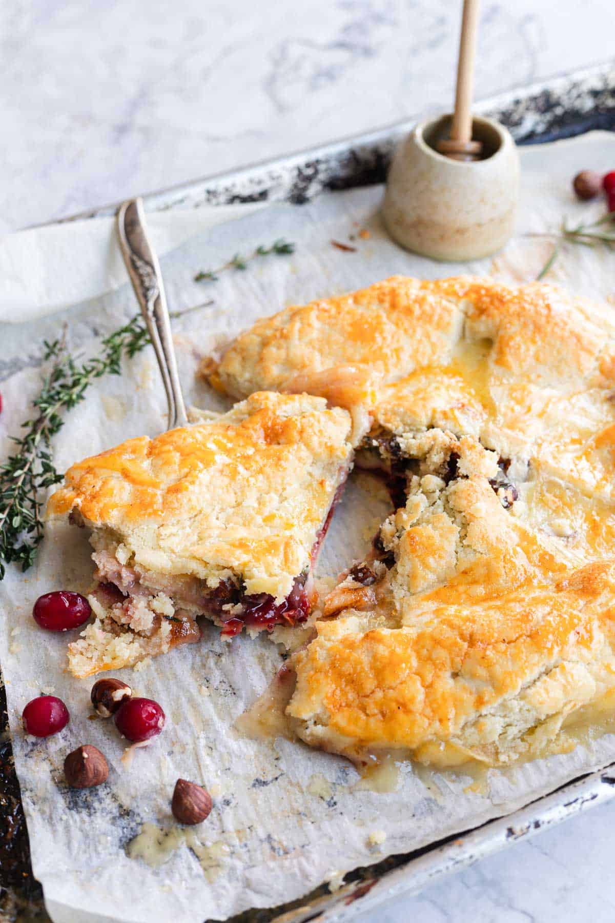 Golden cranberry galette with a slice being served, surrounded by cranberries and herbs on parchment paper&mdash;a rustic twist on a classic Cranberry Brie appetizer.