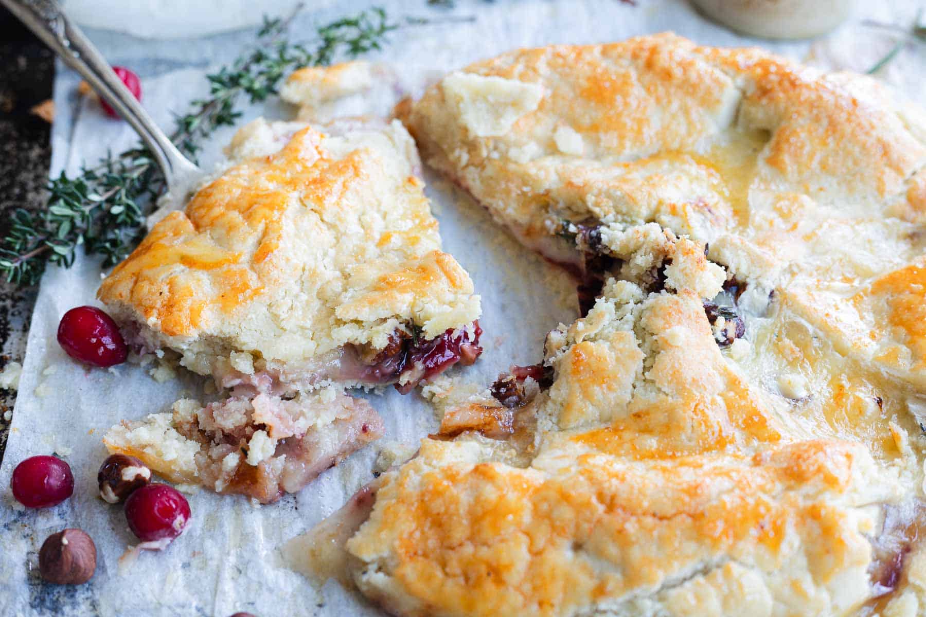 Golden, flaky Cranberry Brie Puff Pastry with fruit filling, sliced open on parchment, surrounded by fresh cranberries and herbs.