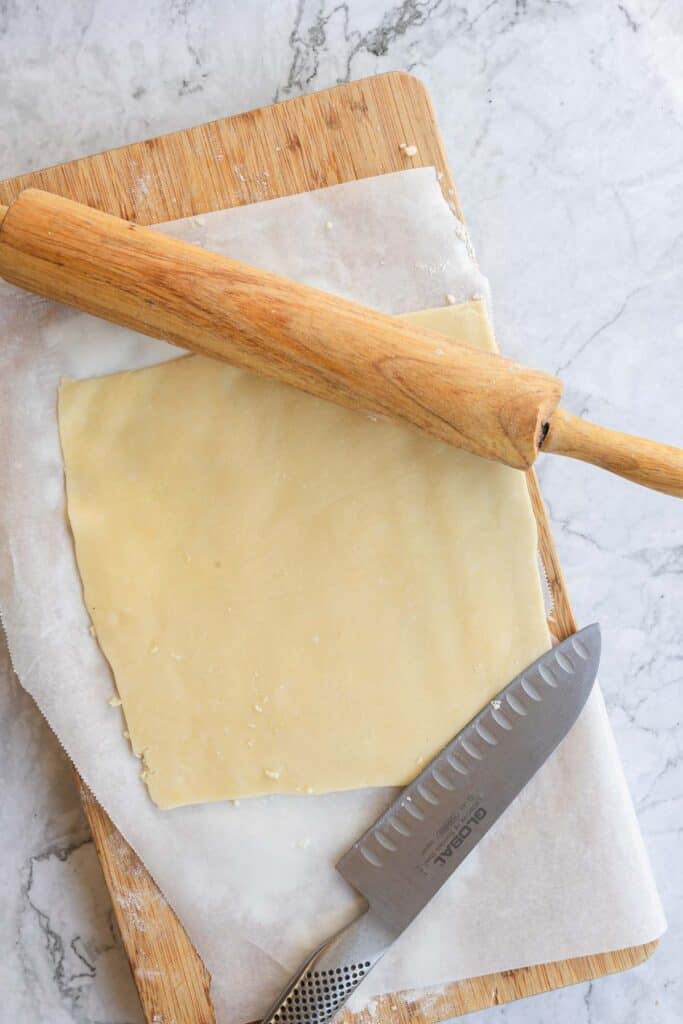 Square puff pastry dough on parchment paper with a rolling pin and knife on a wooden cutting board, ready to be transformed into a delicious Cranberry Brie appetizer.