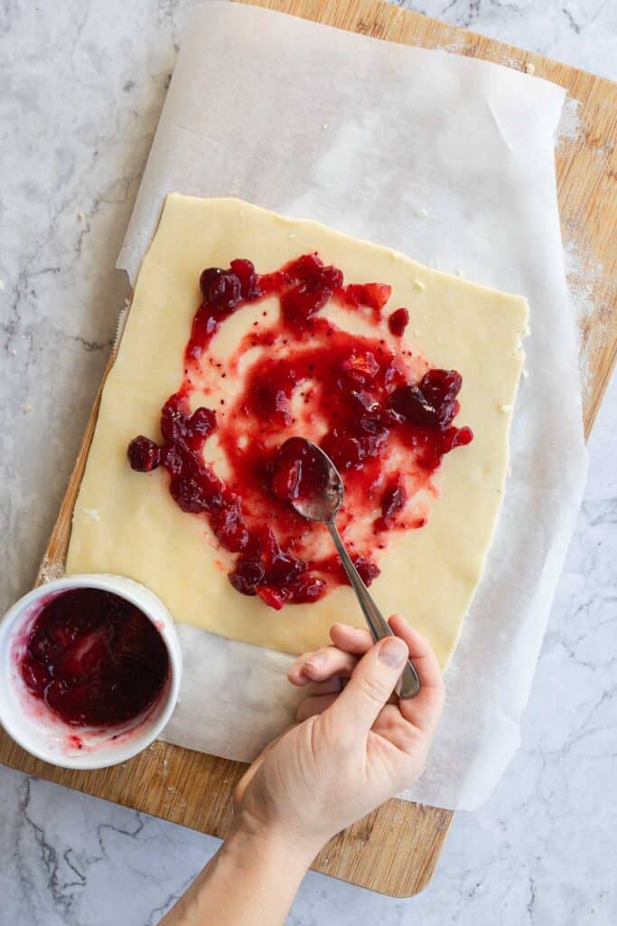 A hand spreading red fruit jam on Puff Pastry dough with a spoon, on parchment paper over a wooden board&mdash;perfect for creating delicious Cranberry Brie Puff Pastry treats.