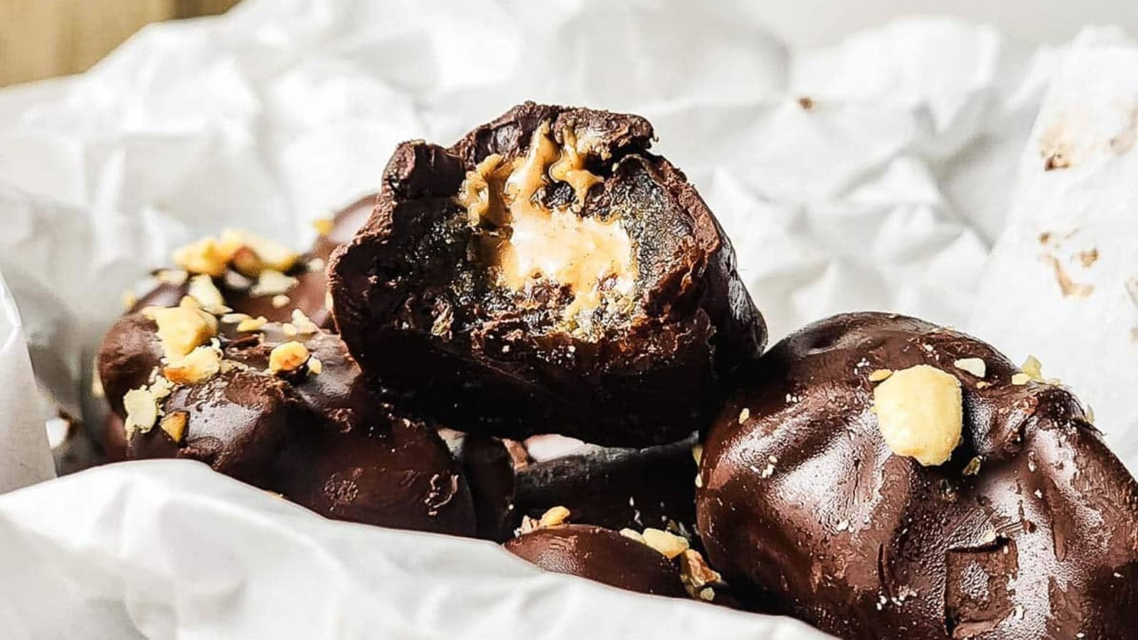 Chocolate-covered candies with a peanut butter filling, topped with crushed nuts, in a box lined with parchment paper.