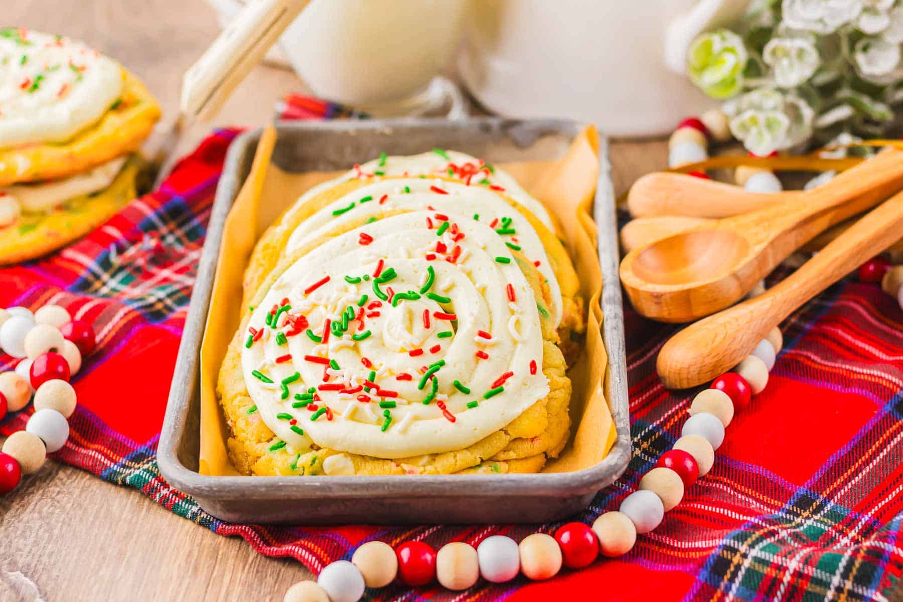 Frosted Christmas cookies made from cake mix, topped with red and green sprinkles, sit in a tray on a plaid cloth with wooden spoons and beads nearby&mdash;a festive holiday recipe display.
