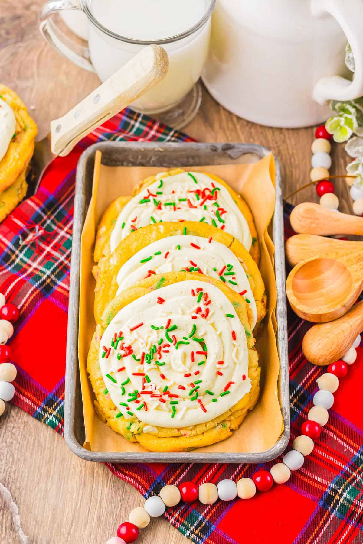 Frosted sugar Christmas cookies made from cake mix with red and green sprinkles in a tray, next to milk on a red plaid cloth&mdash;an easy holiday recipe.