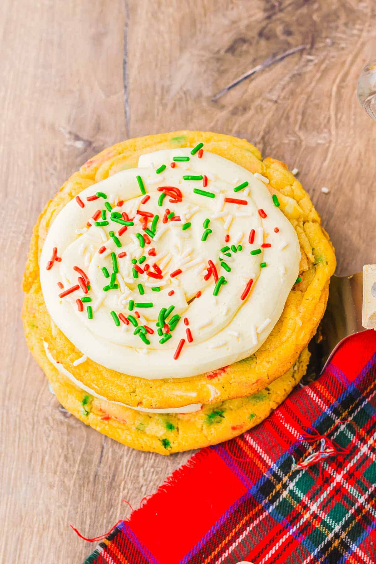 A stack of Christmas cookies made from cake mix, topped with white frosting and red and green sprinkles, sits on a wooden surface by a plaid cloth.