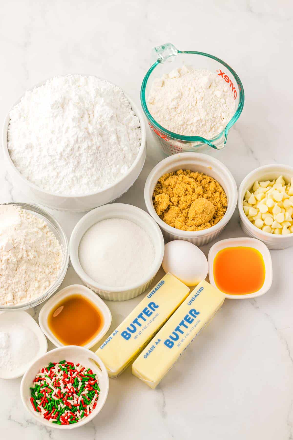 Assorted baking ingredients on a white surface, including flour, sugar, butter, sprinkles, and white chocolate chips&mdash;perfect for your next cake mix or festive Christmas cookies recipe.