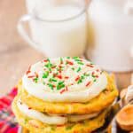 A stack of frosted Christmas cookies with red and green sprinkles, made from a simple cake mix recipe, sits beside a glass of milk in the background.