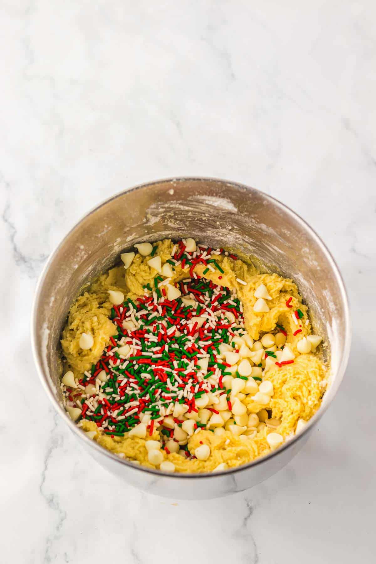 A metal bowl filled with cookie dough, white chocolate chips, and red and green sprinkles on a marble surface&mdash;perfect for your next Christmas cookies recipe.