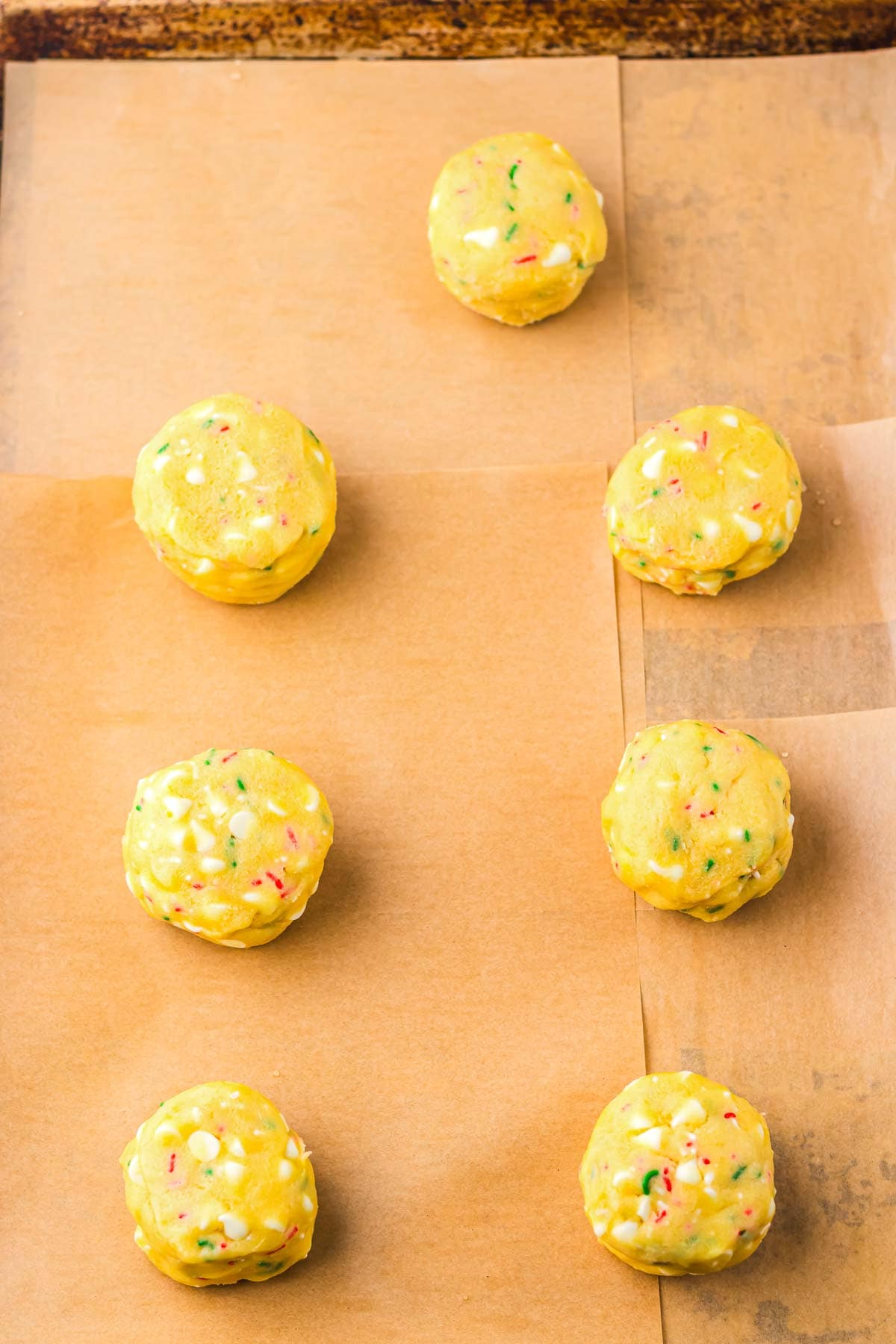 Seven unbaked Christmas Cookies made from cake mix dough sit with sprinkles on parchment paper on a baking sheet, ready for the oven.
