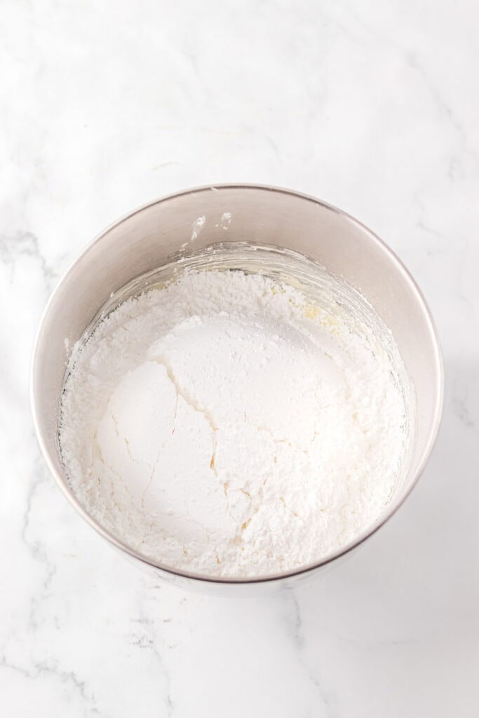 A metal mixing bowl filled with white powdered sugar on a marble countertop, perfect for preparing Christmas cookies.