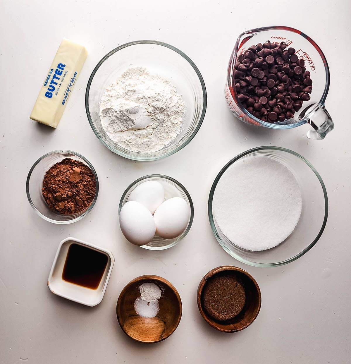 Nine baking ingredients for Fudgy Brownie Cookies, including butter, flour, chocolate chips, eggs, sugar, cocoa, and vanilla, are arranged on a white surface.