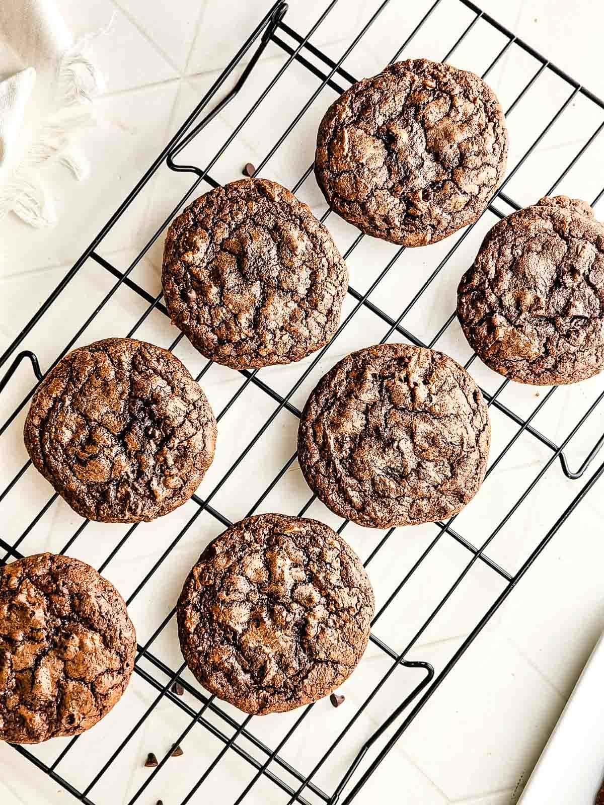 Eight fudgy brownie cookies cooling on a black wire rack placed on a light-colored surface.
