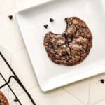A fudgy brownie cookie with a bite taken out, on a white plate, sits next to a cooling rack and scattered chocolate chips.