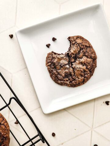 A fudgy brownie cookie with a bite taken out, on a white plate, sits next to a cooling rack and scattered chocolate chips.