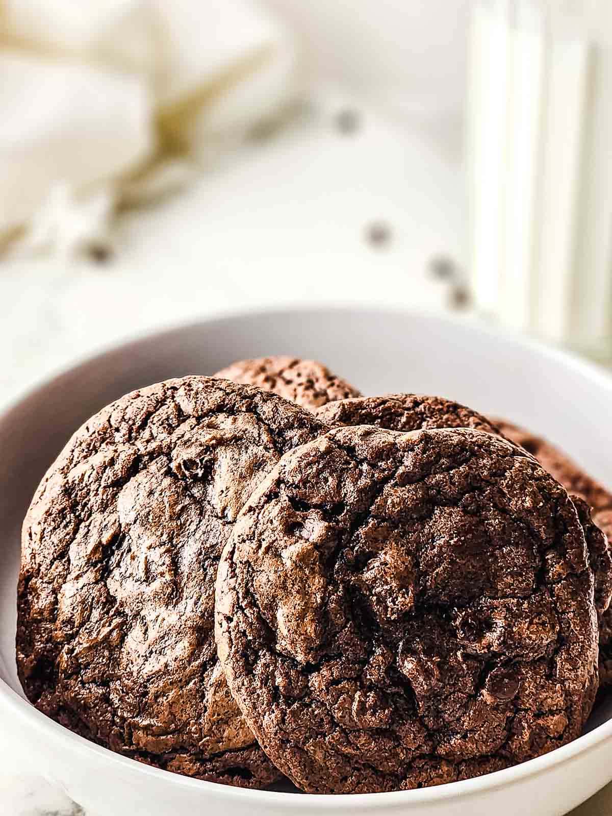 A white bowl filled with fudgy brownie cookies, with a glass of milk blurred in the background.