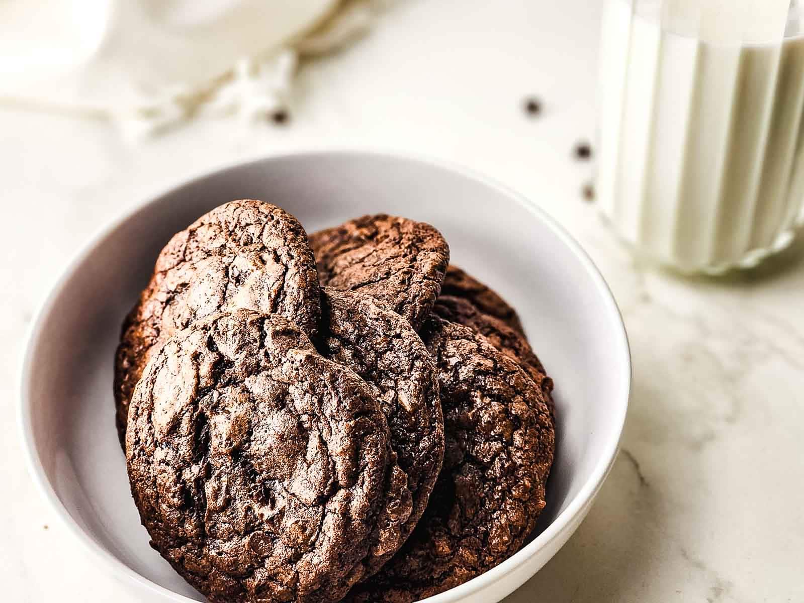 A bowl of fudgy brownie cookies sits next to a glass of milk on a white surface.
