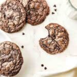 Five fudgy brownie cookies, one with a bite taken out, beside scattered chocolate chips and a glass of milk.