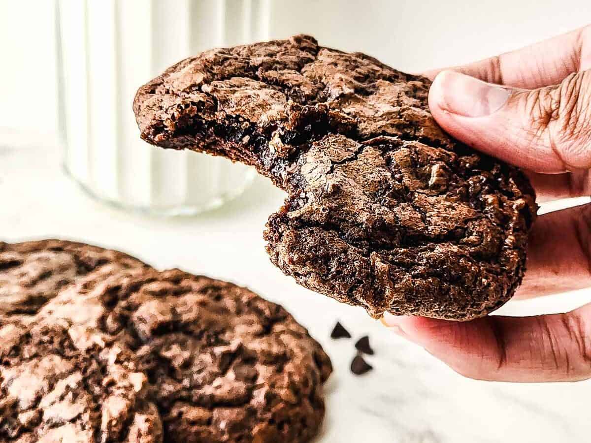 A hand holds a bitten fudgy brownie cookie; more fudgy cookies and a glass of milk are in the background.
